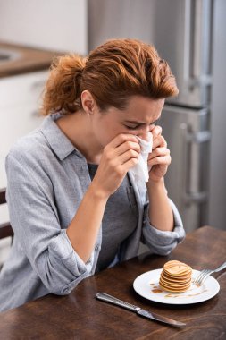 woman sneezing in napkin near tasty pancakes on table  clipart