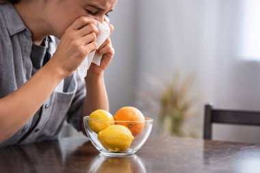 woman with closed eyes sneezing in tissue near orange and lemons in bowl 