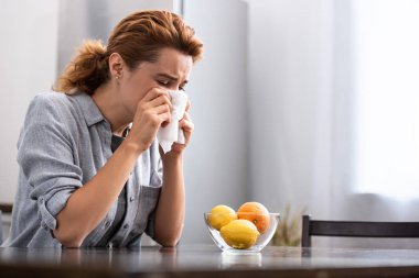 woman with runny nose sneezing in tissue near orange and lemons in bowl  clipart