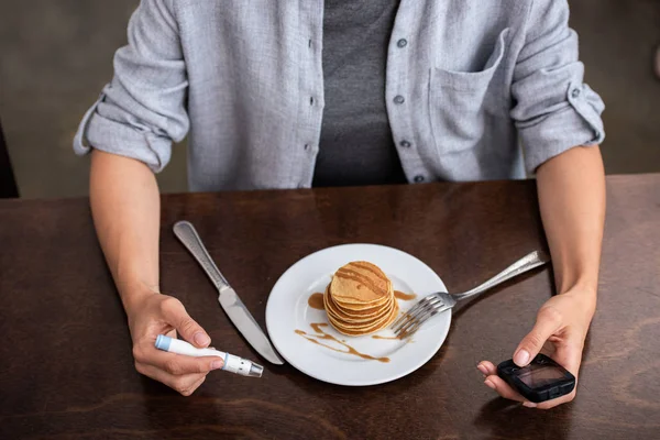 おいしいパンケーキとプレートの近くに血液ランセットとブドウ糖モニターを保持している女性のオーバーヘッドビュー — ストック写真