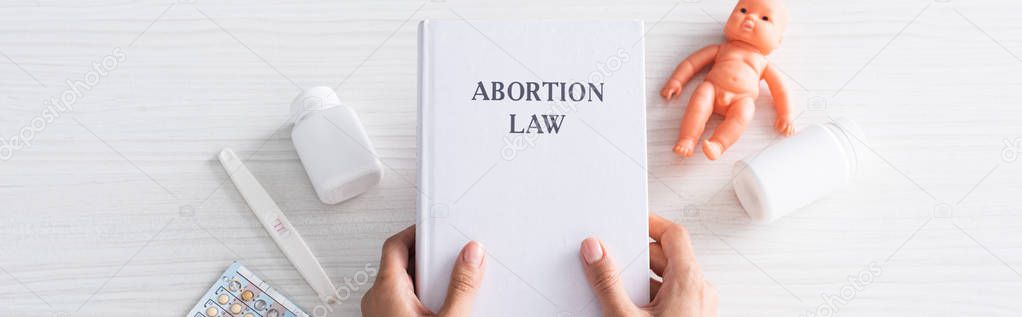 panoramic shot of woman holding book with abortion lettering near baby doll and abortion pills 