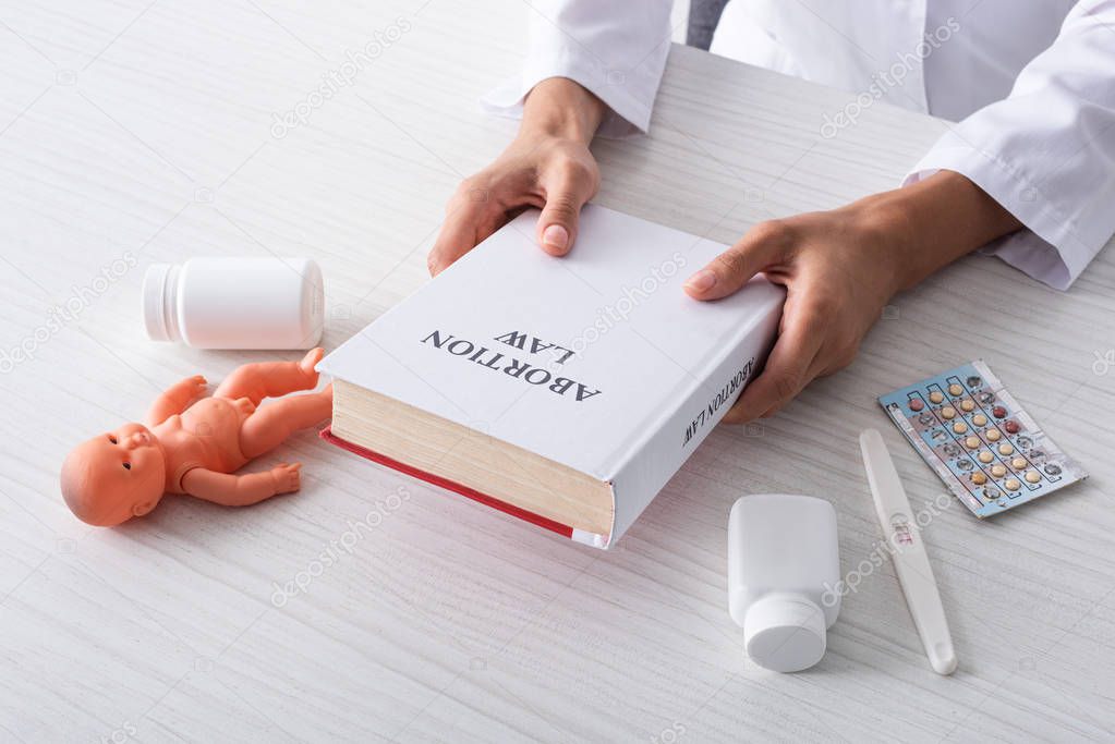 cropped view of doctor holding book with abortion lettering near baby doll and abortion pills 
