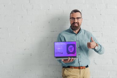 yakışıklı ve gülümseyen işadamı başparmak gösteren ve web-alışveriş web sitesi ile dizüstü tutma 