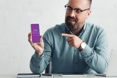 KYIV, UKRAINE - AUGUST 27, 2019: handsome businessman in shirt pointing with finger at smartphone with instagram app