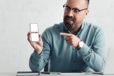 KYIV, UKRAINE - AUGUST 27, 2019: handsome businessman in shirt pointing with finger at smartphone with tinder logo