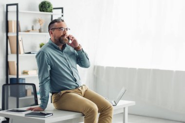 handsome businessman in shirt and glasses sitting on table and talking on smartphone