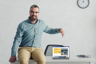 KYIV, UKRAINE - AUGUST 27, 2019: handsome businessman sitting on table and pointing with finger at laptop with booking website 