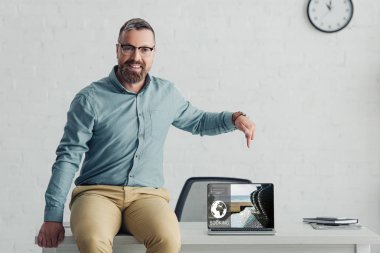 handsome businessman sitting on table and pointing with finger at laptop with booking website 