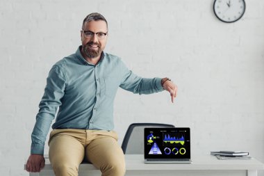 handsome businessman sitting on table and pointing with finger at laptop with charts and graphs website 