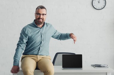 handsome businessman sitting on table and pointing with finger at laptop with copy space