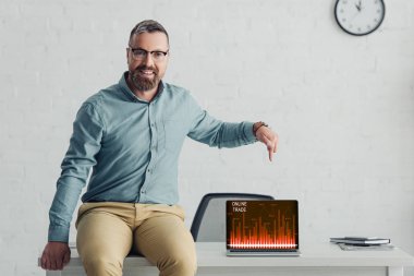 handsome businessman sitting on table and pointing with finger at laptop with online trade website 