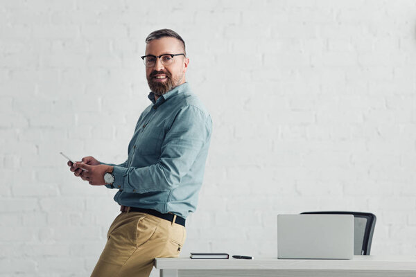 красивый бизнесмен в рубашке, держащий смартфон и смотрящий в камеру
 