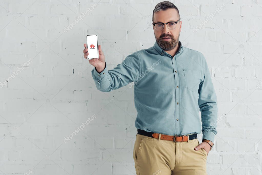 KYIV, UKRAINE - AUGUST 27, 2019: handsome businessman with hand in pocket holding smartphone with huawei logo