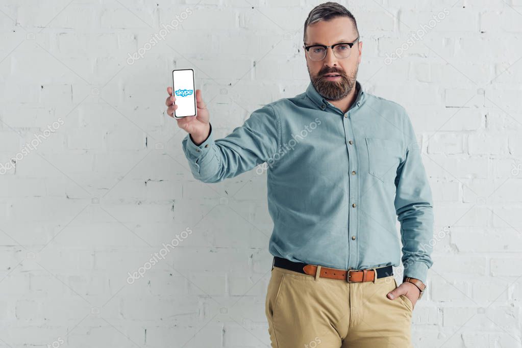 KYIV, UKRAINE - AUGUST 27, 2019: handsome businessman with hand in pocket holding smartphone with skype logo