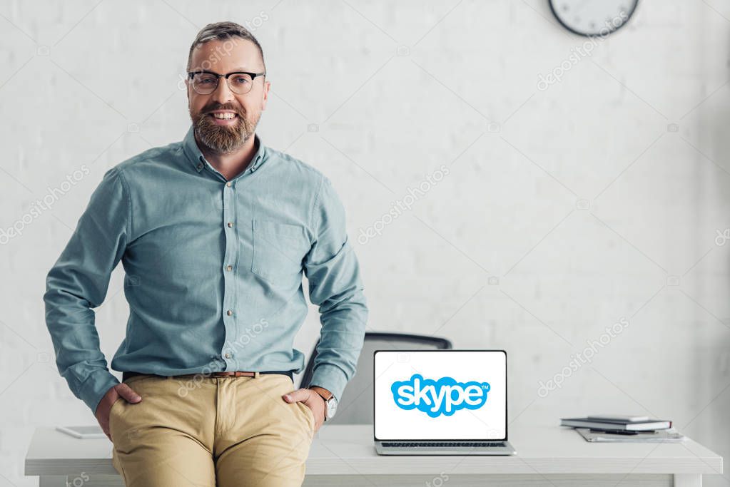 KYIV, UKRAINE - AUGUST 27, 2019: handsome businessman sitting on table near laptop with skype logo
