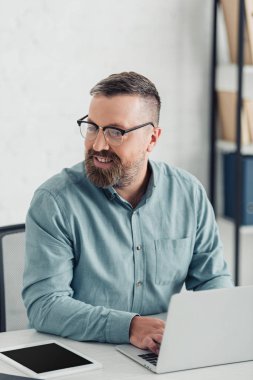 handsome businessman in shirt and glasses using laptop in office  clipart