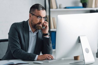 handsome businessman in formal wear talking on smartphone and looking at computer  clipart