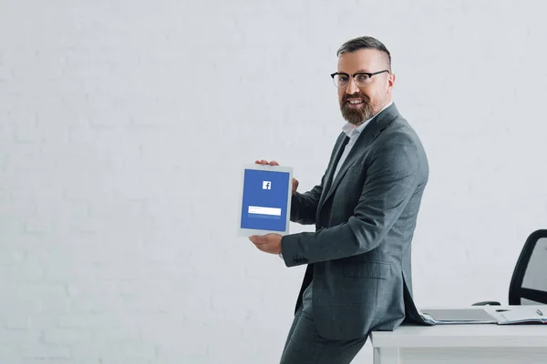 KYIV, UKRAINE - AUGUST 27, 2019: handsome businessman in formal wear holding digital tablet with facebook website