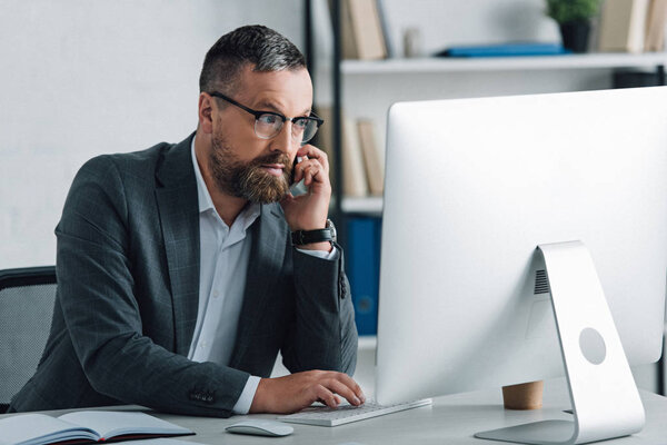 handsome businessman in formal wear talking on smartphone and looking at computer 