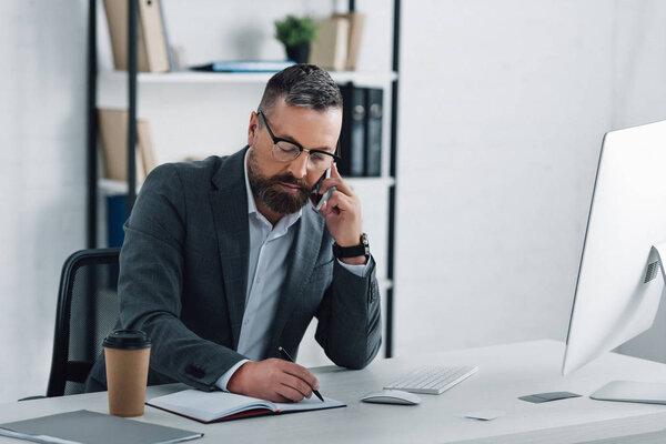 красивый бизнесмен в формальной одежде разговаривает на смартфоне и пишет ручкой
