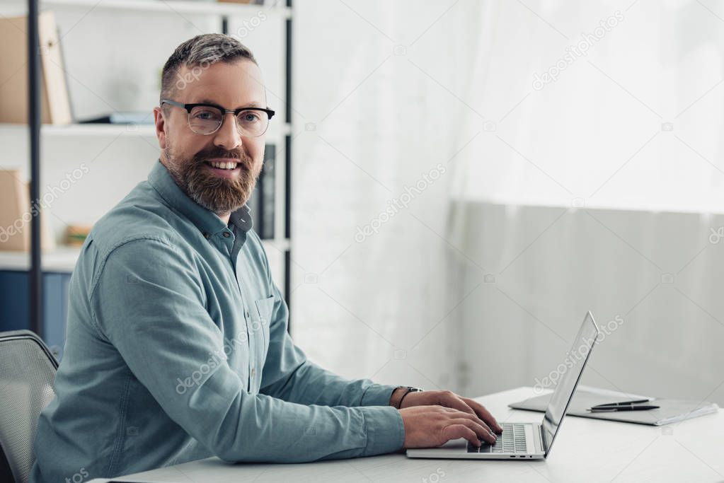 handsome businessman in shirt and glasses using laptop and looking at camera 