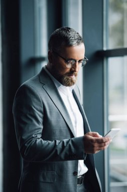 handsome businessman in formal wear and glasses holding smartphone  clipart