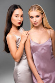 elegant european blonde and asian brunette women in satin dresses isolated on grey