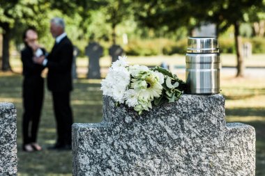 selective focus of bouquet of white flowers and mortuary urn on tombstone near man and woman  clipart