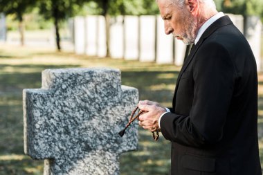man with grey hair looking at rosary beads near tombstone   clipart