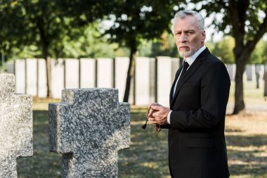 man with grey hair looking at camera and holding rosary beads near tombstones  clipart