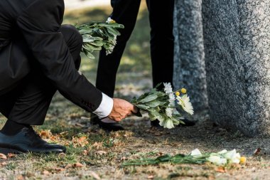 cropped view of senior man putting flowers near tombstones and woman  clipart