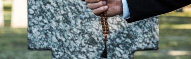 panoramic shot of senior man holding rosary beads near tombstone   clipart