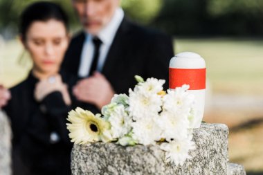 selective focus of white flowers and mortuary urn near man and woman in graveyard  clipart