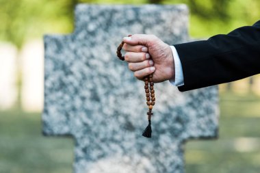 cropped view of elderly man holding rosary beads near gravestone   clipart