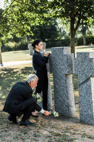 senior man putting flowers near tombs and woman in cemetery 