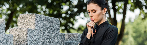 panoramic shot of attractive and sad woman holding rosary beads near tomb 