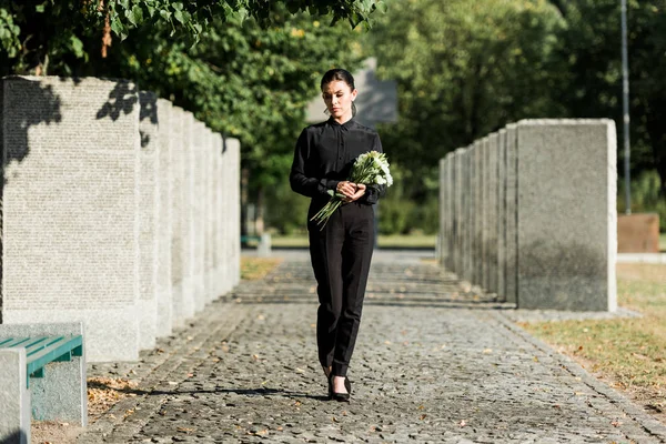 Λυπημένη Γυναίκα Κρατώντας Λουλούδια Και Περπατώντας Στο Νεκροταφείο — Φωτογραφία Αρχείου