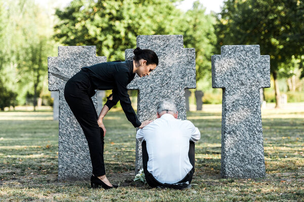 красивая женщина стоит рядом расстроен старший мужчина сидит рядом с могилами
 