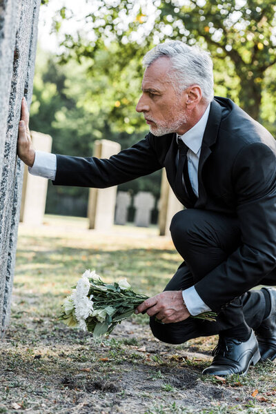 upset man with grey hair putting flowers near tomb in cemetery 