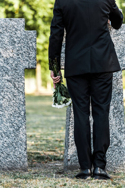 cropped view of senior man holding flowers and standing near tombs 