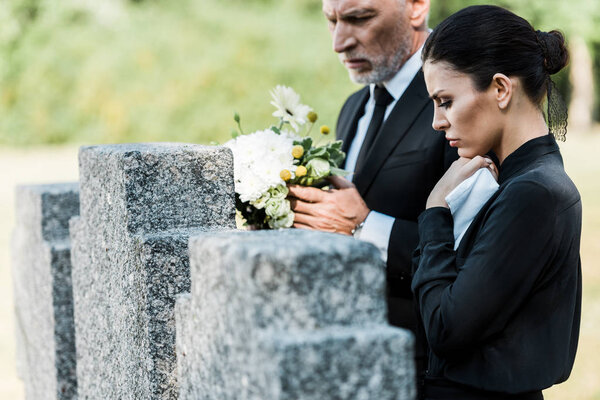 selective focus of man with grey hair holding  flowers near tombstones and woman 