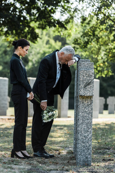 attractive woman holding flowers near tombstones and upset senior man 
