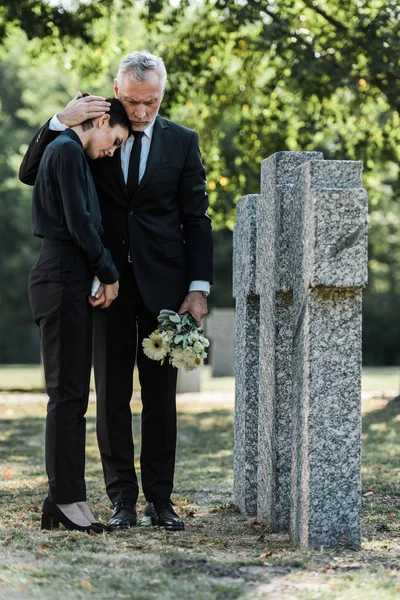 Homem Sênior Segurando Flores Abraçando Mulher Chateada Perto Lápides — Fotografia de Stock