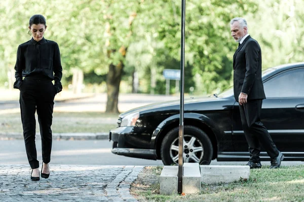 upset man and sad woman in formal wear walking near black car