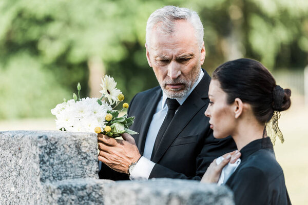 selective focus of upset man holding flowers and standing near woman in cemetery 