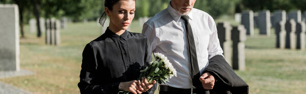 panoramic shot of bearded senior man near woman with flowers on funeral