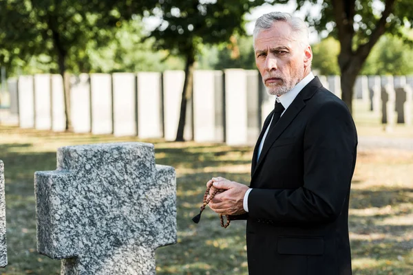 Upprörd Senior Man Innehar Rosenkransen Pärlor Nära Tombstone — Stockfoto