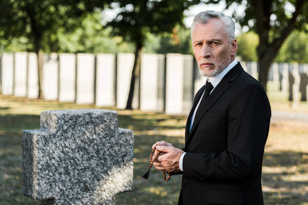 sad man with grey hair holding rosary beads near tombstone  