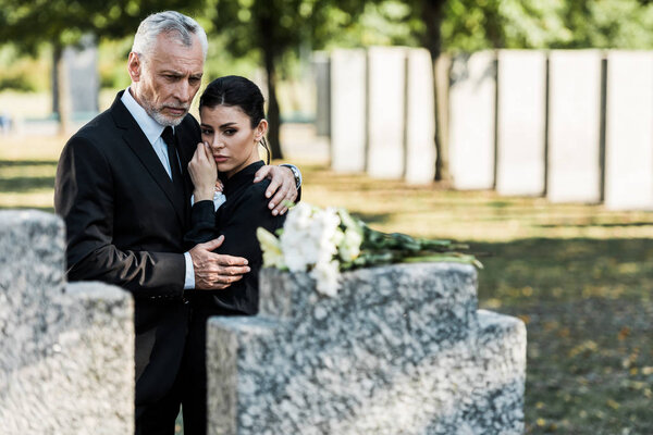 selective focus of man hugging woman while looking at flowers on tomb