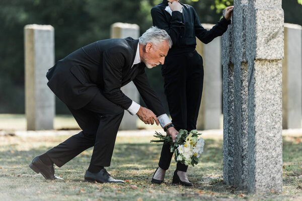 cropped view of woman standing near tomb and bearded man putting flowers on ground 